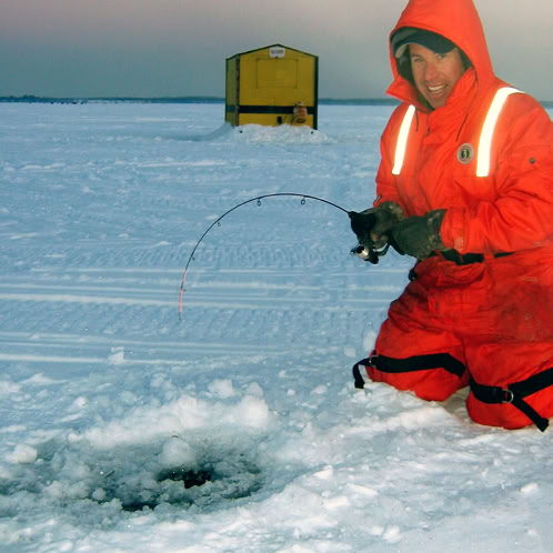 Man in orange snow suit kneeling next to hole in ice catching a fish